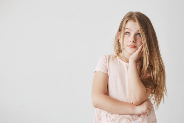 Gros plan d'une belle fille aux cheveux blonds et aux yeux bleus, tenant la main sur la joue, regardant de côté avec une expression rêveuse, pensant aux vacances.