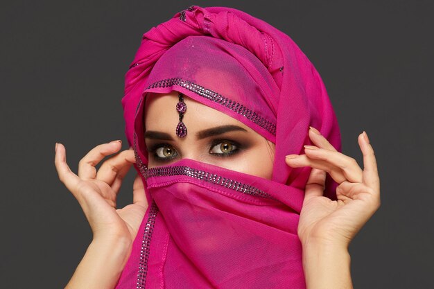 Gros plan d'une belle jeune femme aux yeux charbonneux expressifs portant le hijab rose chic décoré de paillettes et de bijoux. Elle tient le châle avec ses mains et regarde la caméra