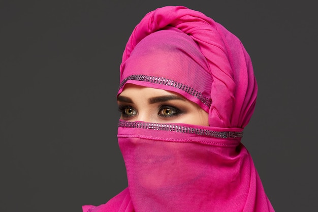 Gros plan d'une belle jeune femme aux yeux charbonneux expressifs portant le hijab rose chic décoré de paillettes. Elle a tourné la tête et détourné les yeux sur un fond sombre. Émot humain