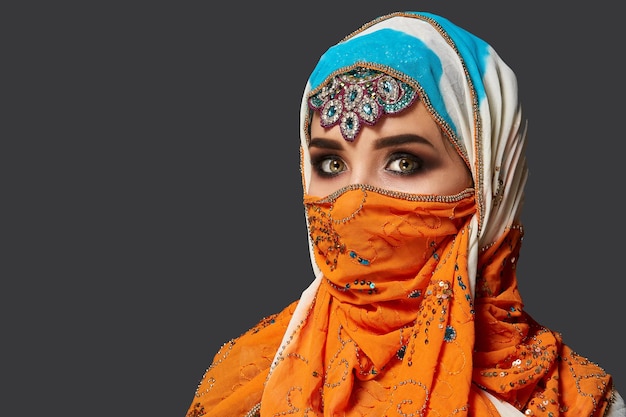 Gros plan d'une belle jeune femme avec un maquillage professionnel portant un élégant hijab coloré décoré de paillettes et de bijoux. Elle pose de côté au studio sur un fond sombre. Émo humain