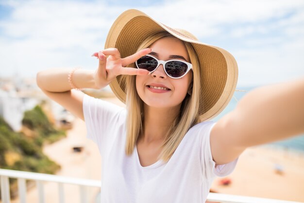 Gros plan d'une belle jeune fille au chapeau d'été prenant un selfie et montrant le geste de paix à la plage