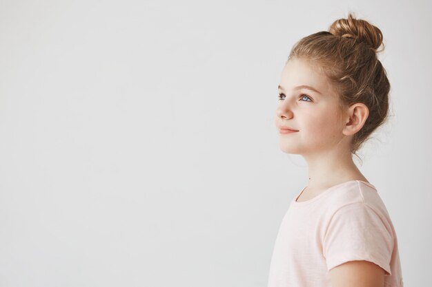 Gros plan d'une belle petite fille aux cheveux blonds en coiffure chignon, debout dans les trois quarts, regardant de côté avec le sourire sur son visage.