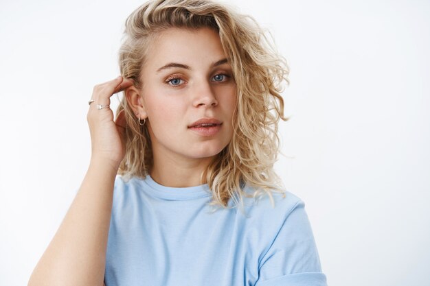 Gros plan d'une blonde douce sensuelle et féminine avec une coiffure courte bouclée mettant une mèche de cheveux derrière l'oreille bouche ouverte avec un regard séduisant et romantique, posant sur un mur blanc