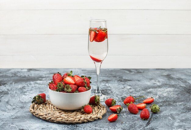 Gros plan un bol de fraises sur napperon rond en osier avec un verre de boisson sur marbre bleu foncé et surface de planche de bois blanc. horizontal