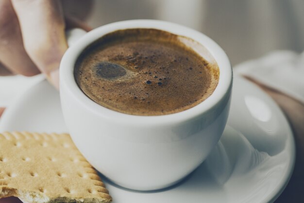 Gros plan d&#39;un café expresso savoureux avec de la mousse savoureuse dans une petite tasse en céramique. Main femelle tenant une boisson chaude chaude.