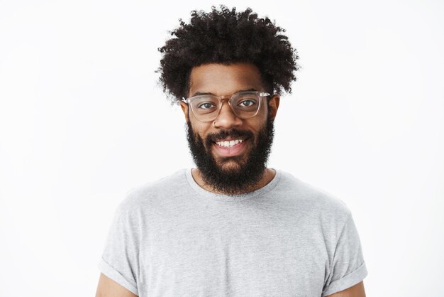 Gros plan sur un charmant client masculin barbu à la peau sombre, agréable et ravi, dans des verres avec une coiffure afro en t-shirt décontracté gris, souriant joyeusement comme ayant une bonne humeur sur le gris.