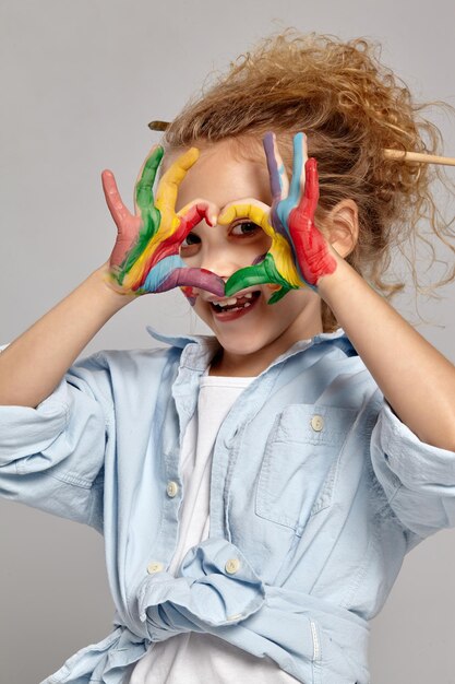 Gros plan d'un charmant enfant aux joues peintes ayant un pinceau dans ses cheveux blonds bouclés chics, portant une chemise bleue et un t-shirt blanc. Elle regarde à travers un bras peint plié dans le shap