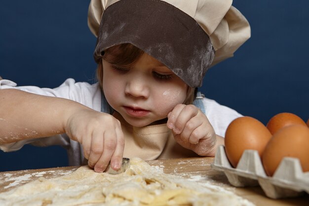 Gros plan de la charmante petite fille mignonne portant un grand chapeau de chef faisant des biscuits à la table de la cuisine, à l'aide de moules de boulangerie, ayant concentré l'expression concentrée. Enfants, concept de cuisine et de pâtisserie