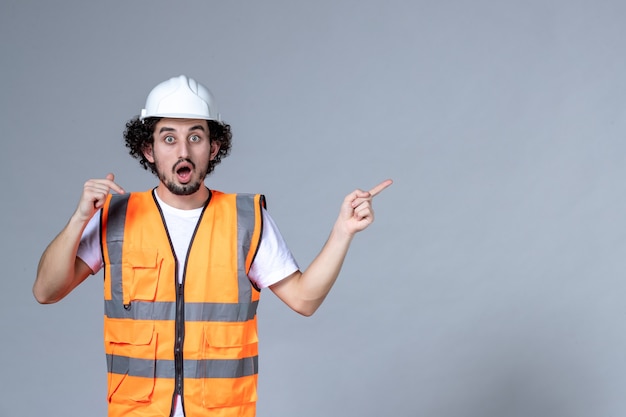 Gros plan d'un constructeur masculin choqué dans un gilet d'avertissement portant un casque de sécurité et pointant quelque chose sur le côté gauche sur le mur de la vague grise