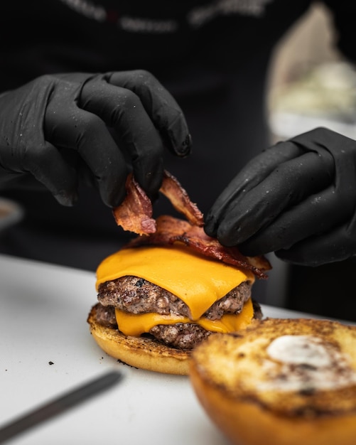Gros plan d'un cuisinier plaçant du bacon frit sur une tranche de fromage tout en superposant un hamburger