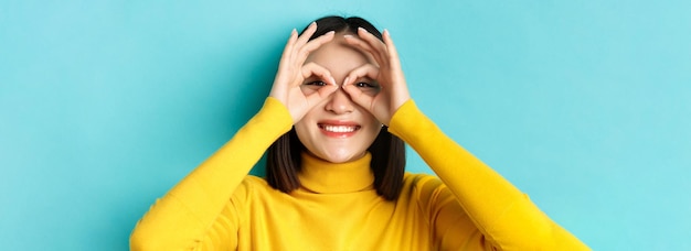 Photo gratuite gros plan d'une drôle de fille asiatique regardant à travers des lunettes à main et souriant vérifiant le stand de l'offre promotionnelle