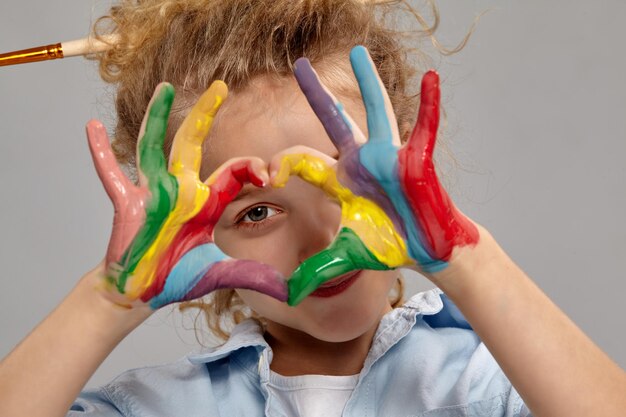 Gros plan d'une drôle de petite fille ayant une brosse dans ses cheveux blonds bouclés chics, vêtue d'une chemise bleue et d'un t-shirt blanc. Elle regarde à travers une mains peintes pliées en forme de coeur et s