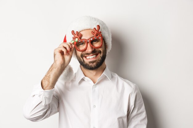 Gros plan du beau mec barbu dans des lunettes de fête de Noël et bonnet de Noel, souriant et souhaitant joyeux Noël, debout