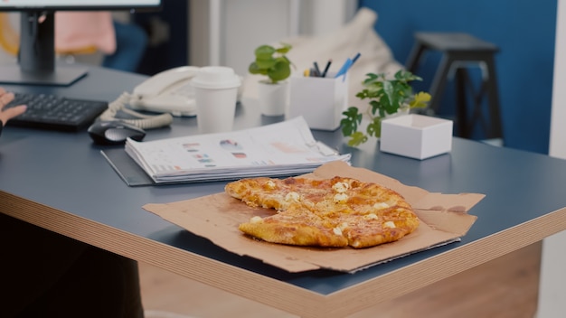 Gros plan du directeur exécutif prenant une tranche de pizza en train de manger devant un ordinateur en train de taper des graphiques financiers dans le bureau de l'entreprise en démarrage