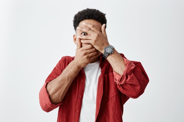 Gros plan du jeune homme à la peau foncée avec une coiffure afro en e-shirt blanc sous le visage de vêtements de chemise rouge avec les mains, regardant à travers les doigts avec des expressions de visage effrayé.
