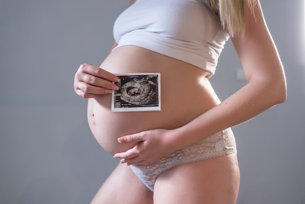 Gros plan du ventre de jeune modèle gravé montrant l'image ultrasonore de son bébé. Future maman sur son deuxième trimestre tenant l'échographie de son enfant. Concept de maternité