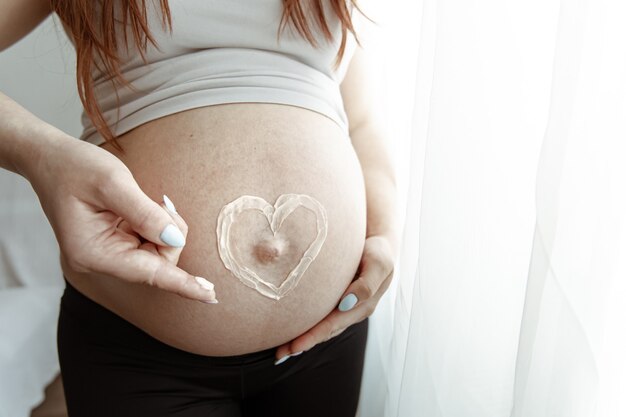 Gros plan du ventre nu d'une future maman dans les derniers mois de grossesse avec une crème coeur peinte.