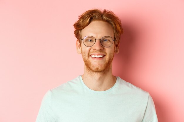 Gros plan du visage de l'homme rousse heureux, souriant avec des dents blanches à la caméra, portant des lunettes pour une meilleure vue et un t-shirt, debout sur fond rose.