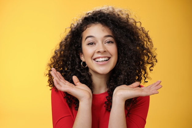 Gros plan d'une étudiante gentille et tendre, jolie caucasienne aux cheveux bouclés et à la peau parfaite souriante ravie tenant les paumes écartées près du visage s'amusant sur fond jaune.