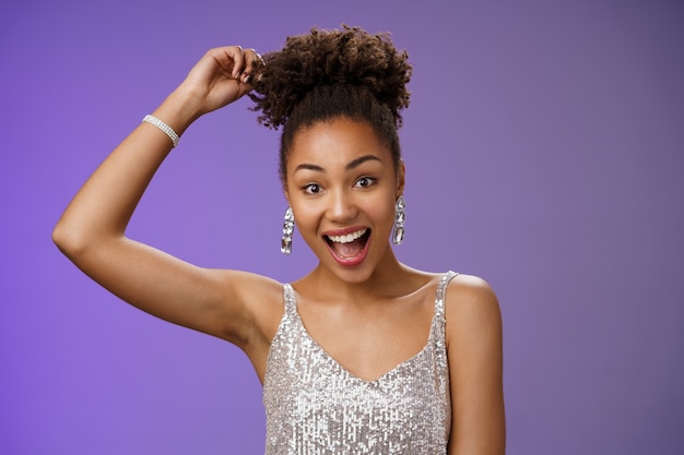 Gros plan excité belle femme afro-américaine aux cheveux bouclés en robe scintillante argentée à la mode touchant afro bouclé souriant joyeusement s'amusant à danser amusé debout amusé fond bleu.