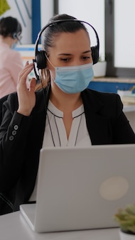 Gros plan sur une femme d'affaires avec un masque facial portant un casque parlant dans un microphone tout en tapant des statistiques marketing sur un ordinateur portable. indépendant assis au bureau au bureau pendant la pandémie de covid19