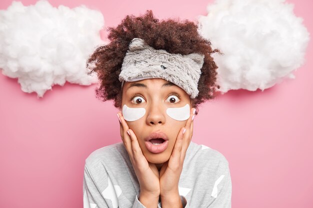 Gros plan d'une femme afro-américaine bouclée choquée garde les mains sur les joues ouvre la bouche à regarde la caméra porte des patchs de collagène bandés sous les yeux pose en pyjama se prépare à dormir