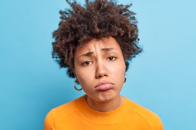 Gros plan d'une femme afro-américaine bouleversée avec une expression piteuse, les lèvres des sacs à main ont une expression triste et triste exprime des émotions négatives pose contre le mur bleu offensé