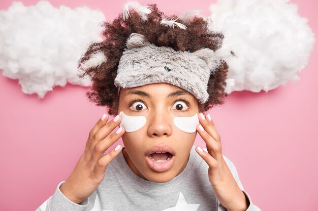 Gros plan d'une femme afro-américaine émotive étonnée applique des patchs sous les yeux regarde la caméra porte un bandeau doux subit des procédures de soins de la peau pose sur les nuages de mur rose au-dessus