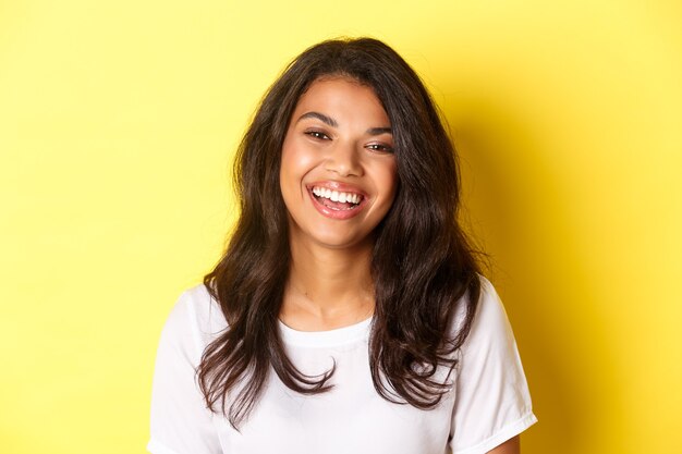 Gros plan d'une femme afro-américaine joyeuse et belle, riant et ayant l'air heureux, debout sur fond jaune.