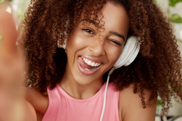 Gros plan d'une femme afro-américaine joyeuse écoute une musique agréable avec des écouteurs, pose pour selfie, être de bonne humeur. Adolescente à la peau sombre enteratins elle-même avec un appareil moderne