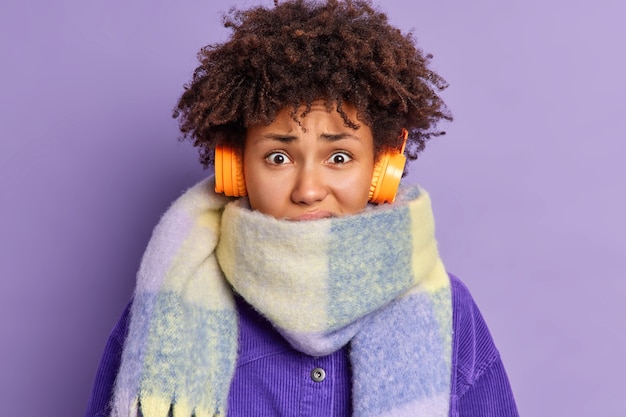 Photo gratuite gros plan d'une femme afro-américaine mécontente de frissons de froid porte une écharpe chaude autour du cou passe beaucoup de temps à l'extérieur pendant l'hiver écoute la piste audio via des écouteurs.