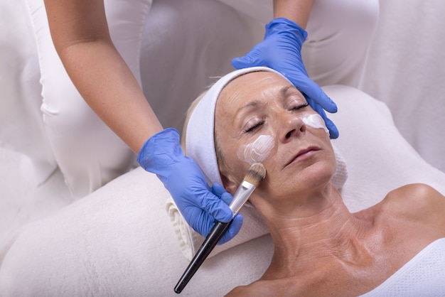 Gros plan d'une femme âgée de race blanche appliquant une crème pour le visage dans un salon de beauté