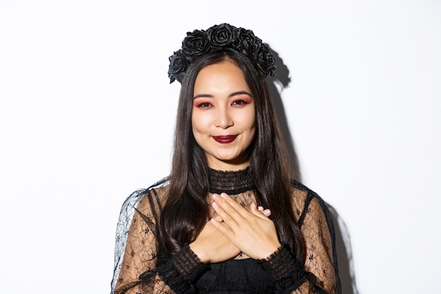 Gros plan d'une femme asiatique souriante reconnaissante à la reconnaissance avec les mains sur la poitrine, debout en costume de sorcière sur fond blanc.