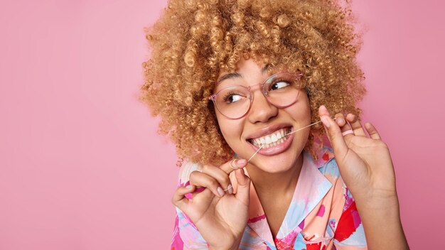 Gros plan d'une femme aux cheveux bouclés uss fil dentaire prend soin de l'hygiène bucco-dentaire regarde au loin porte des lunettes transparentes pose sur fond rose copiez l'espace pour votre contenu promotionnel