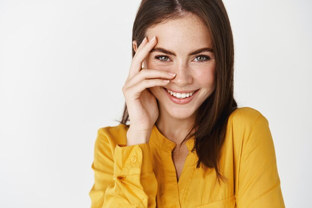 Gros plan d'une femme belle et romantique regardant le flirt devant, souriant et touchant une peau propre et éclatante, semblant en bonne santé et heureuse, debout sur un mur blanc.