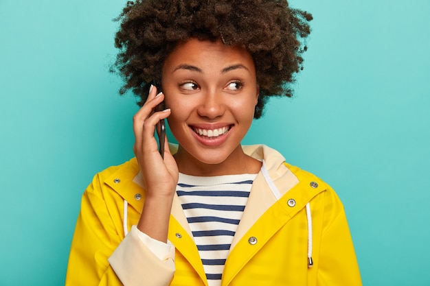 Gros plan d'une femme bouclée a une expression de visage heureux, appelle son meilleur ami pour raconter une histoire hilarante, vêtu d'un imperméable jaune imperméable, concentré sur le côté, se tient à l'intérieur sur fond bleu.