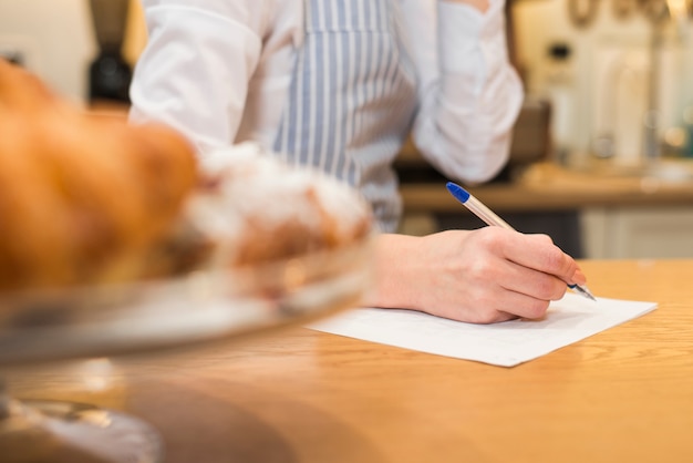 Gros plan, femme, boulanger, écriture, papier blanc, stylo, dessus, table bois