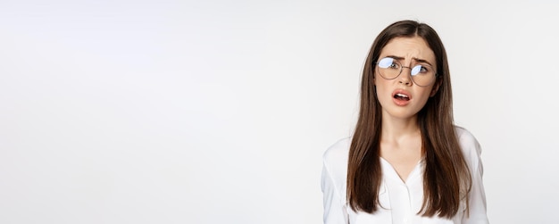 Gros plan sur une femme brune confuse dans des verres qui a l'air perplexe et désemparé debout sur le dos blanc