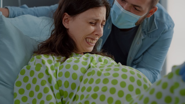 Gros plan sur une femme enceinte souffrant de douleur lors de l'accouchement d'un enfant dans un lit d'hôpital. Homme caucasien aidant sa femme avec les contractions du travail, donnant de l'aide. Couple en couches se préparant à la parentalité