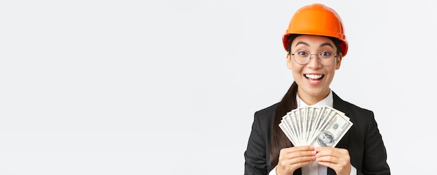 Gros plan d'une femme entrepreneure asiatique heureuse et excitée à l'usine portant un casque de sécurité et un costume d'affaires montrant de gros revenus détenant de l'argent et se réjouissant souriant fond blanc heureux