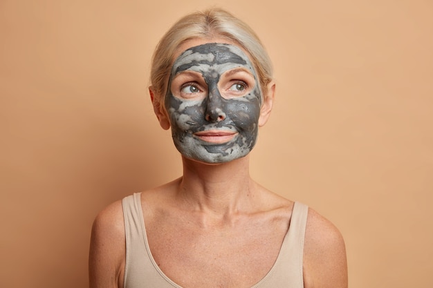 Gros plan d'une femme européenne réfléchie applique un masque d'argile du visage et regarde ailleurs se soucie du teint pose avec les épaules nues isolé sur mur marron