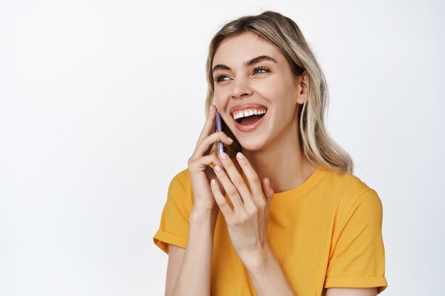 Gros plan d'une femme heureuse parlant au téléphone portable, riant sur appel, debout sur un t-shirt jaune blanc.