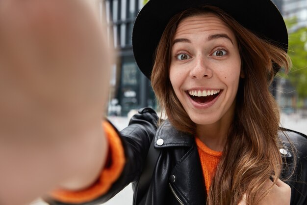 Gros plan d'une femme heureuse porte un chapeau noir, sourit largement, étant de bonne humeur, s'amuse pendant la promenade à l'extérieur