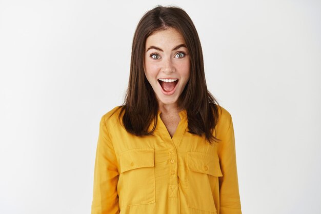 Gros plan sur une femme heureuse et surprise qui ouvre la bouche et regarde avec incrédulité, reçoit de bonnes nouvelles, exprime son étonnement en se tenant debout sur un mur blanc.