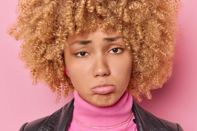 Gros plan d'une femme malheureuse et frustrée qui porte les lèvres et regarde tristement la caméra a une expression faciale déprimée pose contre le mur rose du studio se sent triste. Concept d'émotions humaines négatives