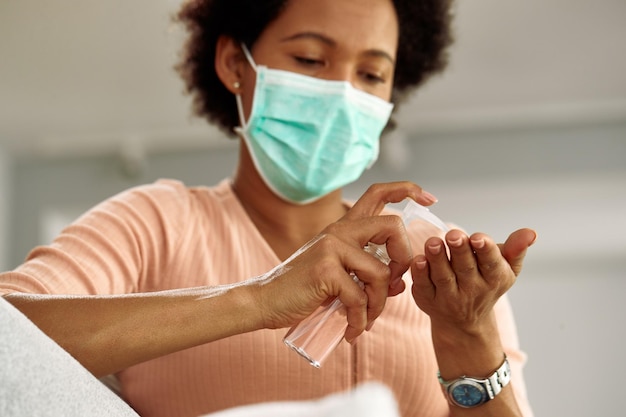 Gros plan d'une femme noire avec un masque facial utilisant un gel antiseptique tout en se nettoyant les mains à la maison