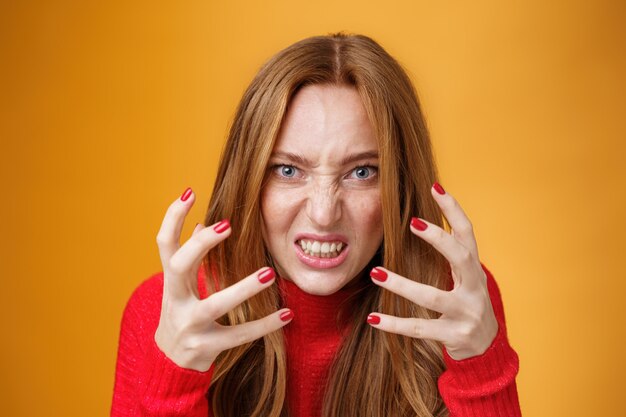 Gros plan d'une femme rousse énervée et en colère levant les mains et les serrant avec colère et indignation, irritée et agacée en grimaçant de l'aversion, de la haine et de la pression sur le mur orange