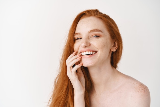 Gros plan d'une femme rousse heureuse à la peau pâle et parfaite, riant et montrant des dents blanches, debout nue sur le mur du studio