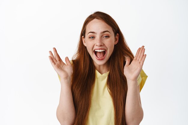 Gros plan d'une femme rousse heureuse qui rit, applaudit et sourit étonnée, vérifiant l'offre promotionnelle impressionnante, bonne nouvelle, debout sur fond blanc.