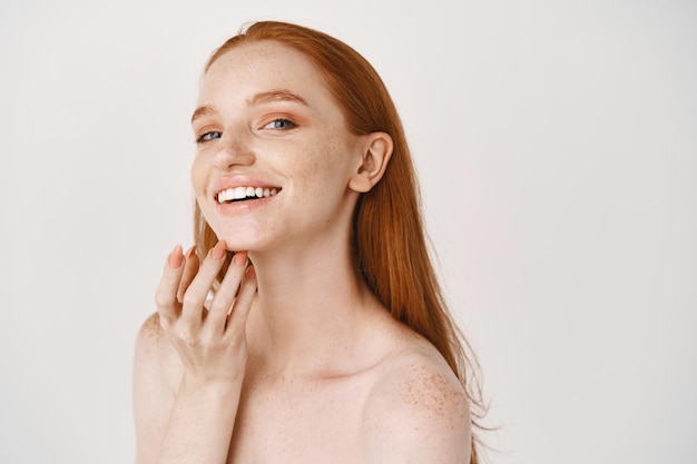 Gros plan d'une femme rousse souriante à la peau pâle et aux taches de rousseur touchant un visage doux et parfait, utilisant une crème de soin, debout sur un mur blanc